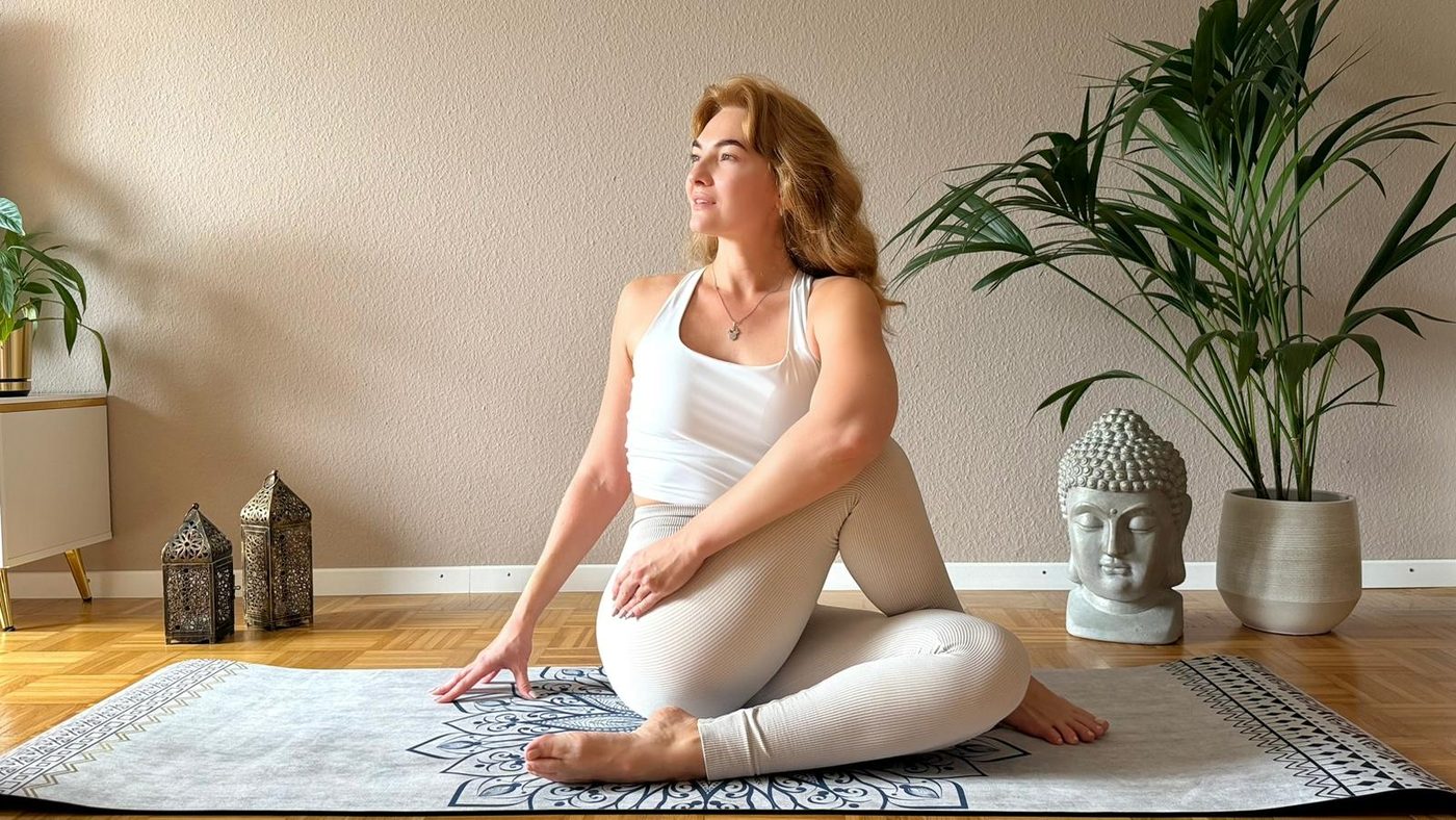 Anastasiia Mamlina sitzt in einer Drehhaltung auf der Yogamatte in einem hellen Wohnraum